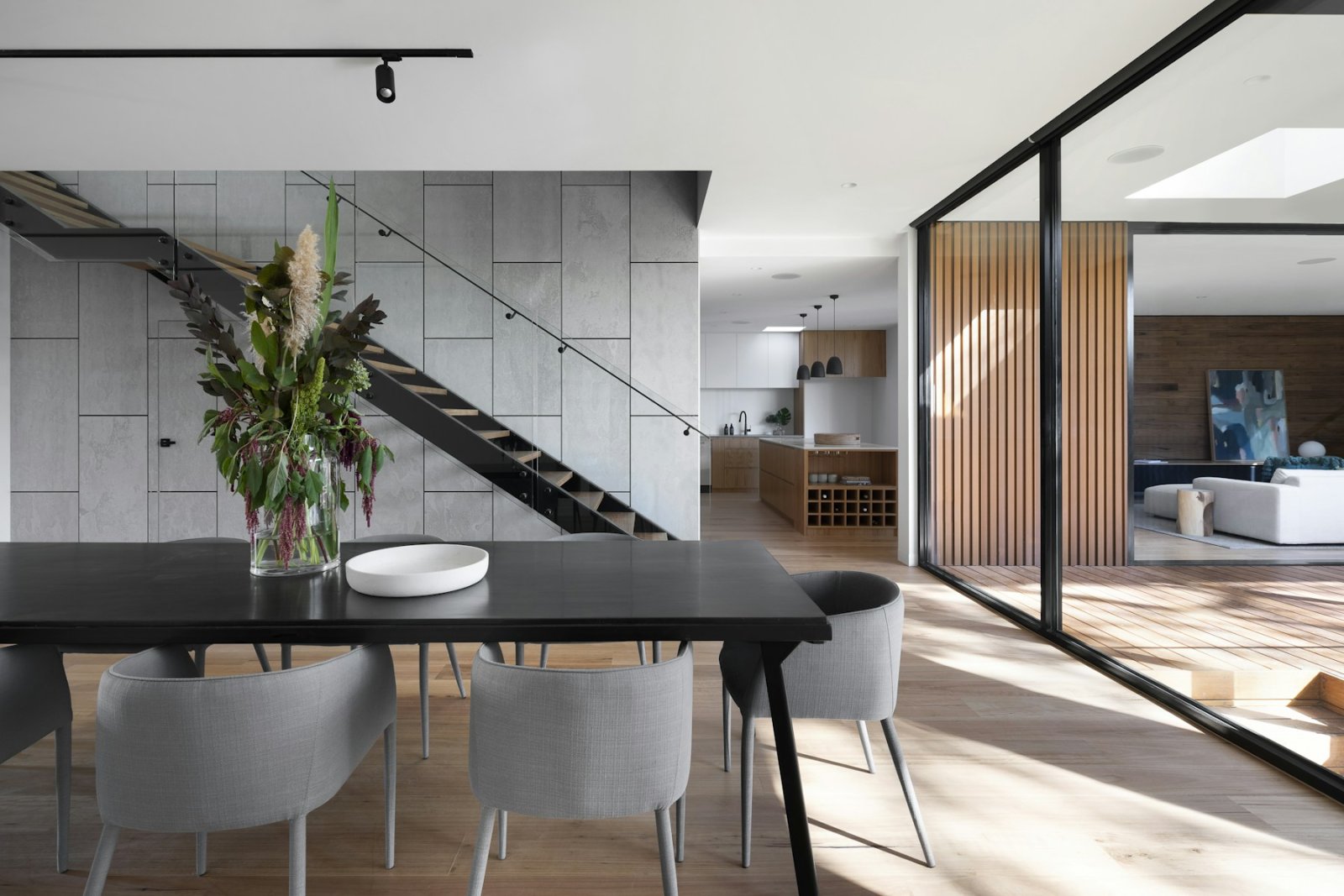 Dining area with a black table and grey chairs beside a concrete-look wall, a steel staircase and glazed sliding doors