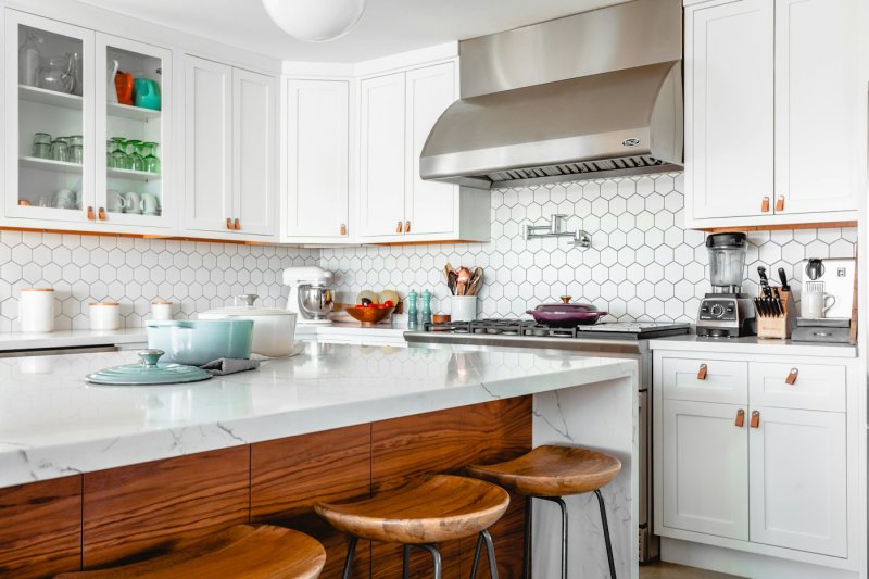 White kitchen with a marble island, timber-clad base, hexagonal tile splashback and timber bar stools