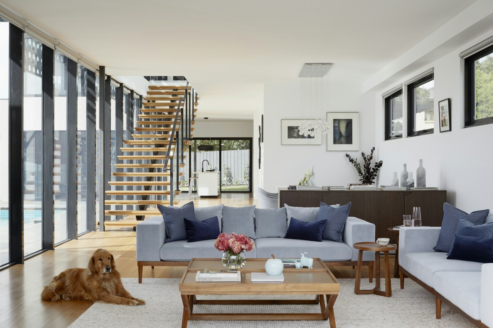 Open-plan living room with a floating timber staircase, soft grey sofas, a timber coffee table and a golden retriever